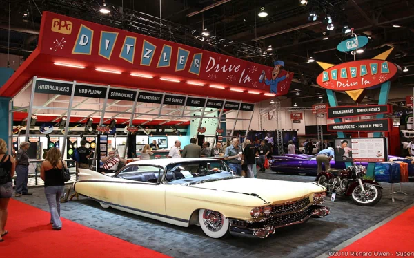 HD PC desktop wallpaper: cream vintage Cadillac displayed at an indoor car show beneath retro Ditzler Drive-In signage, surrounded by other classic cars and attendees.