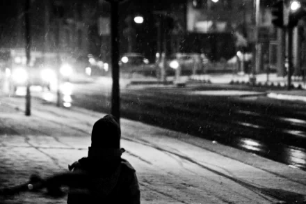 A monochrome scene captures a figure in a winter setting, with snow falling softly, illuminated by distant headlights on a quiet street, showcasing a contrast of dark and light.