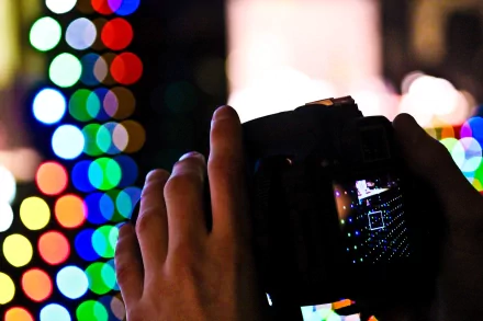 A close-up of a hand holding a Canon camera capturing bright, colorful bokeh lights against a dark background, presented as a 4K Ultra HD desktop wallpaper.