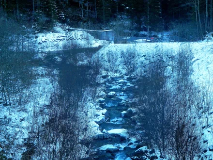 A serene winter landscape photograph featuring a snow-covered forest and a flowing river, captured in stunning 4K Ultra HD for a nature-themed PC desktop wallpaper.