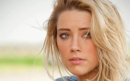 A close-up of Amber Heard with tousled blonde hair and captivating blue eyes, set against a soft, natural background, designed as an HD desktop wallpaper.