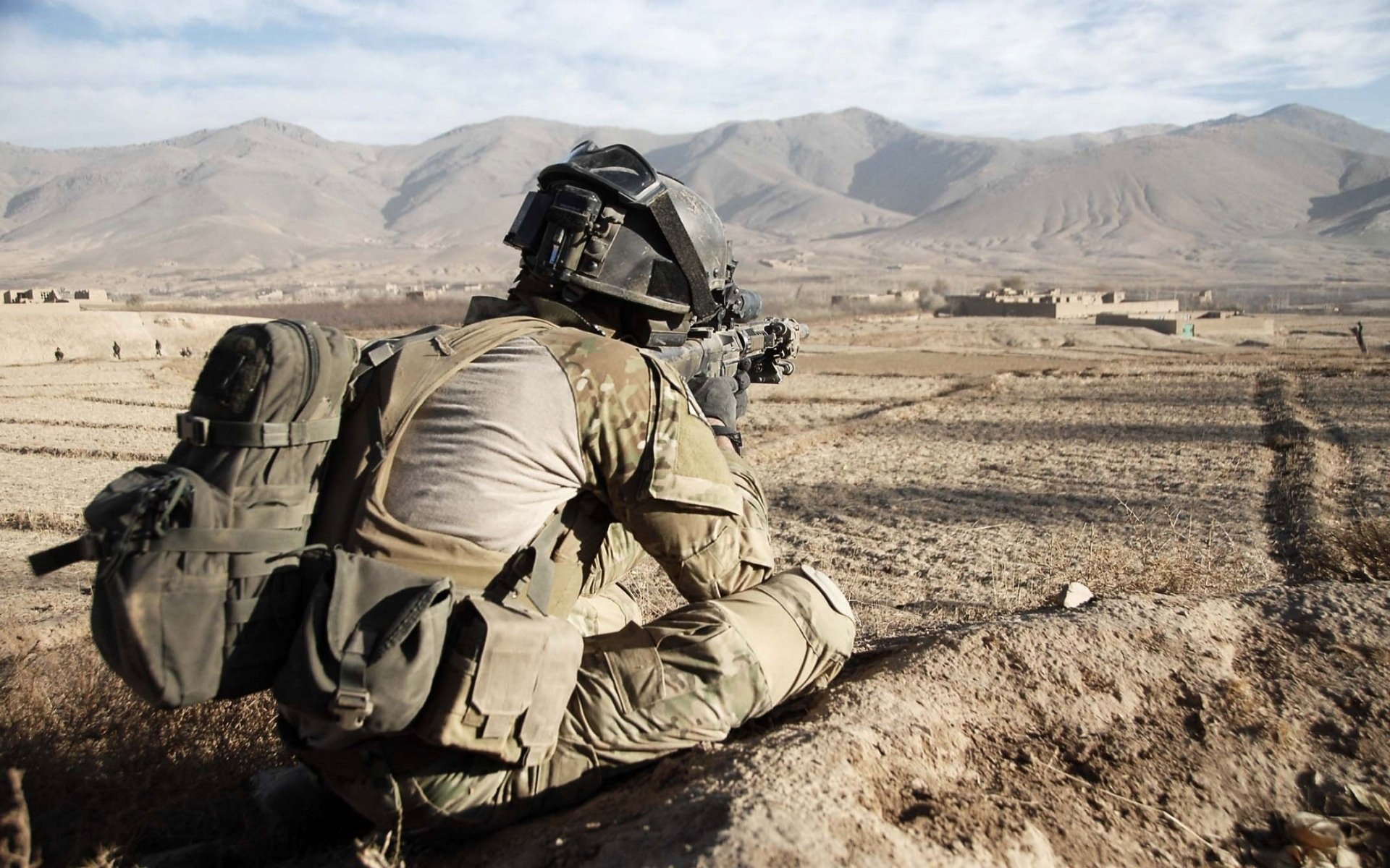 HD PC desktop wallpaper: soldier in tactical gear lying prone in an arid landscape, aiming a rifle across a dusty plain toward distant mountains.