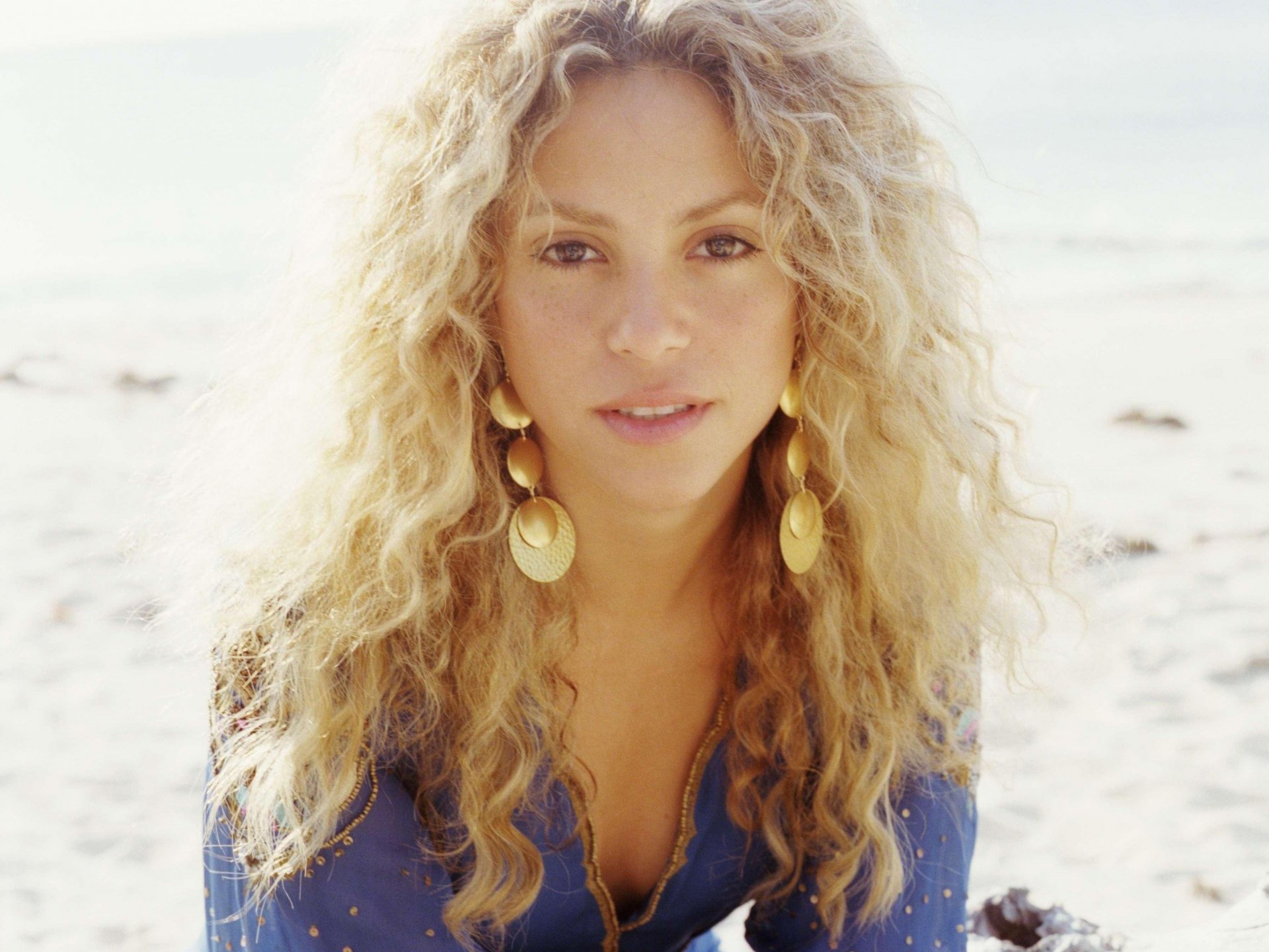 2K Quad HD PC wallpaper: close-up portrait of a music artist with long curly blonde hair and large gold earrings, bathed in soft sunlight on a sandy beach.