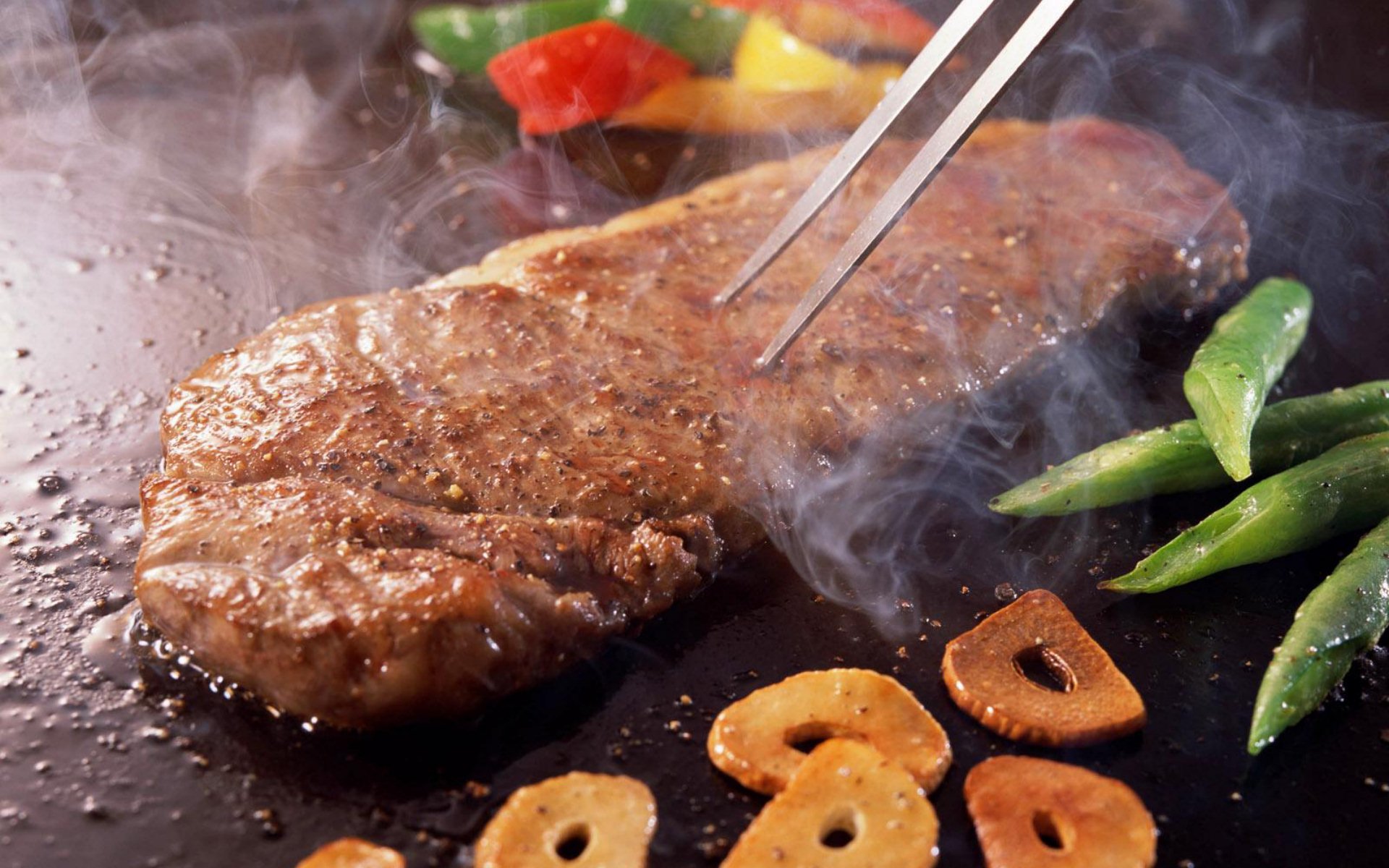HD PC desktop wallpaper showing a sizzling barbecue steak with grilled vegetables and garlic slices on a smoky grill.