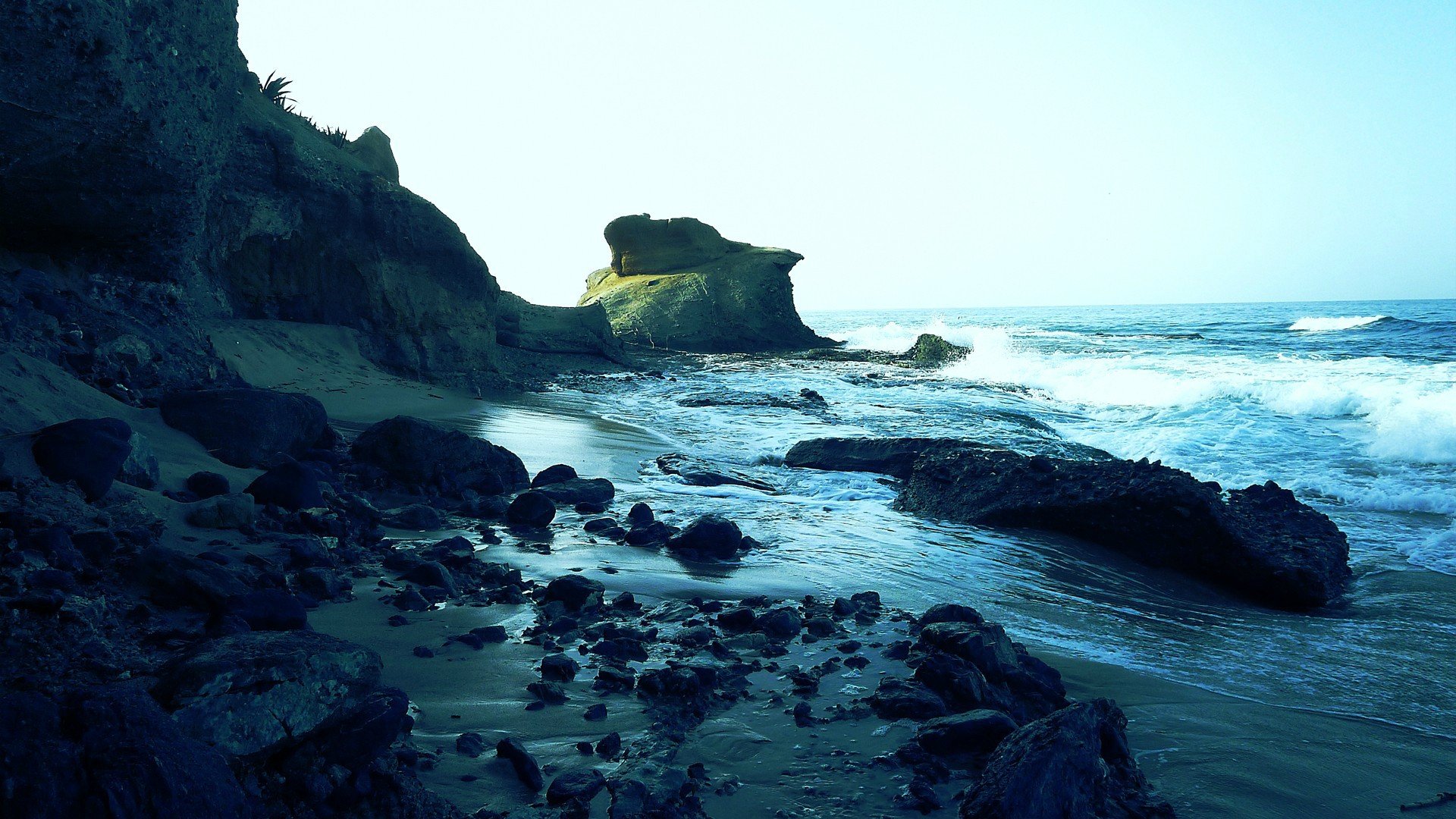 HD PC desktop wallpaper featuring a serene beach scene with rocky shoreline, calm water, and natural coastal landscape under bright daylight.