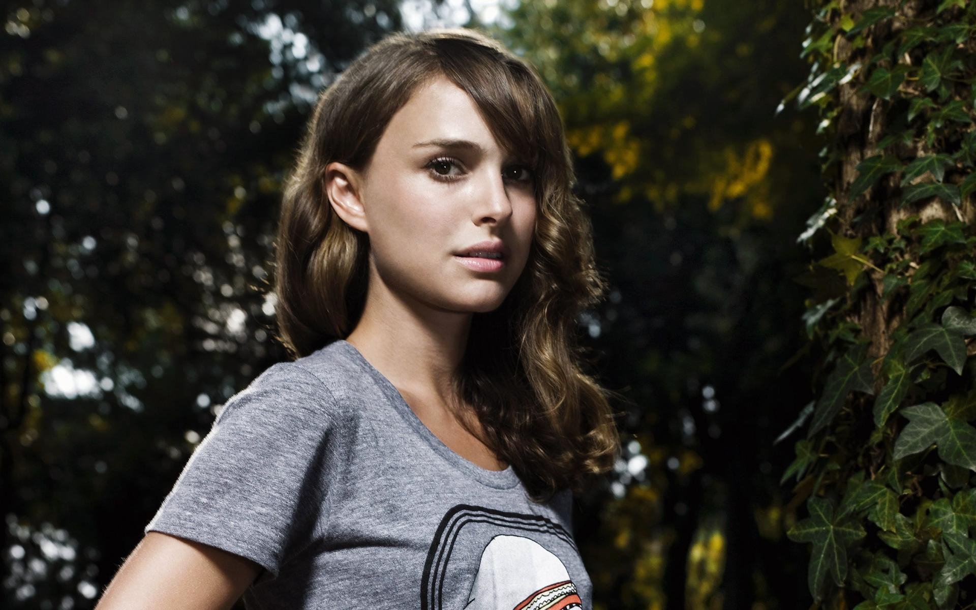 HD desktop wallpaper featuring celebrity Natalie Portman in a casual grey t-shirt, standing outdoors with trees and greenery in the background.