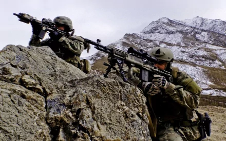 HD desktop wallpaper showing U.S. Army Infantry soldiers in military gear aiming weapons behind rocky terrain in a mountainous war zone.