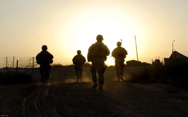 HD desktop wallpaper: silhouettes of four U.S. Army infantry soldiers walking down a dusty road at sunset in a war-torn landscape, military gear and rifles visible against a glowing sky.