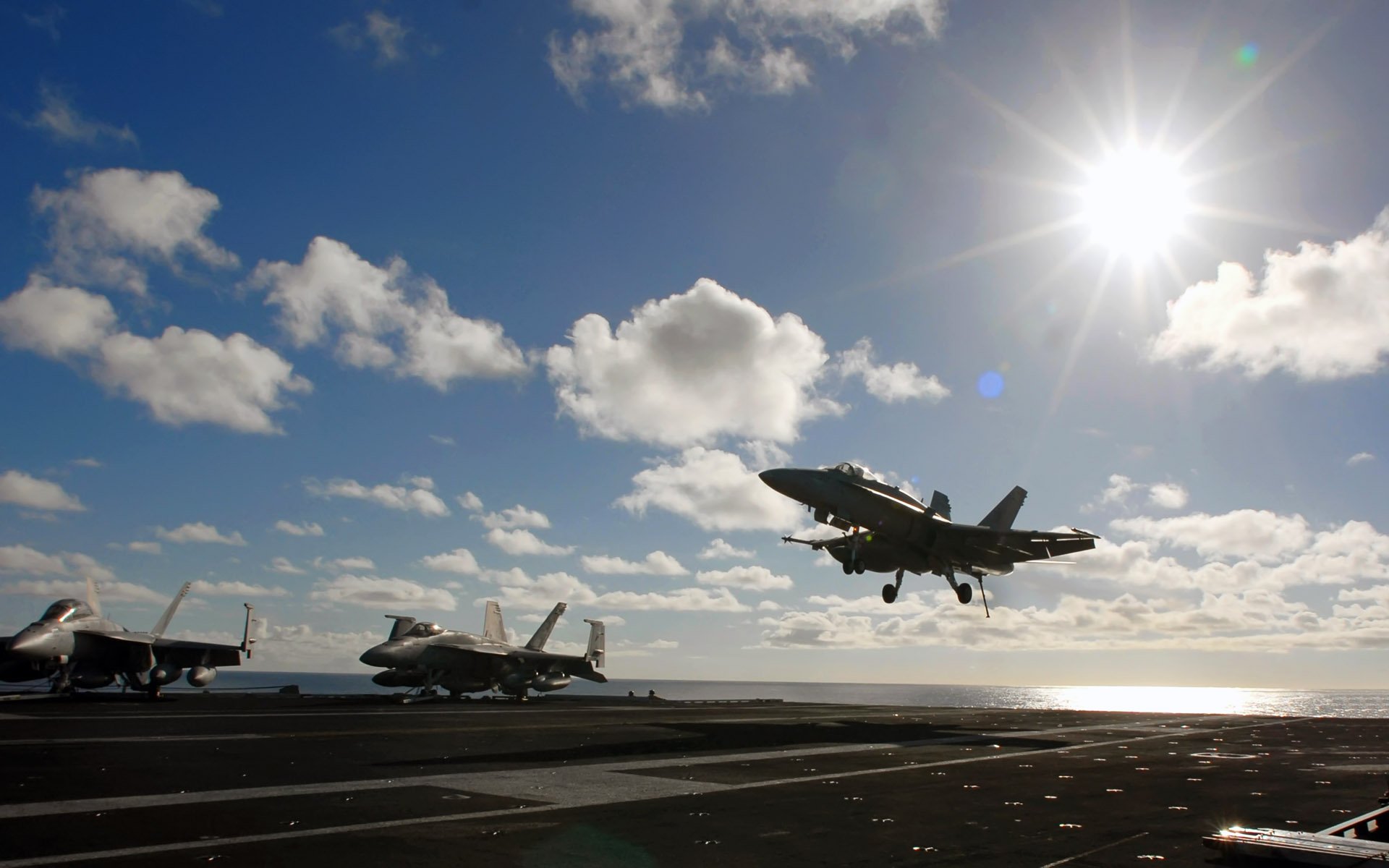 McDonnell Douglas F/A-18 Hornet military aircraft on a carrier deck with one jet landing under a bright sun and scattered clouds, captured in HD for desktop wallpaper.
