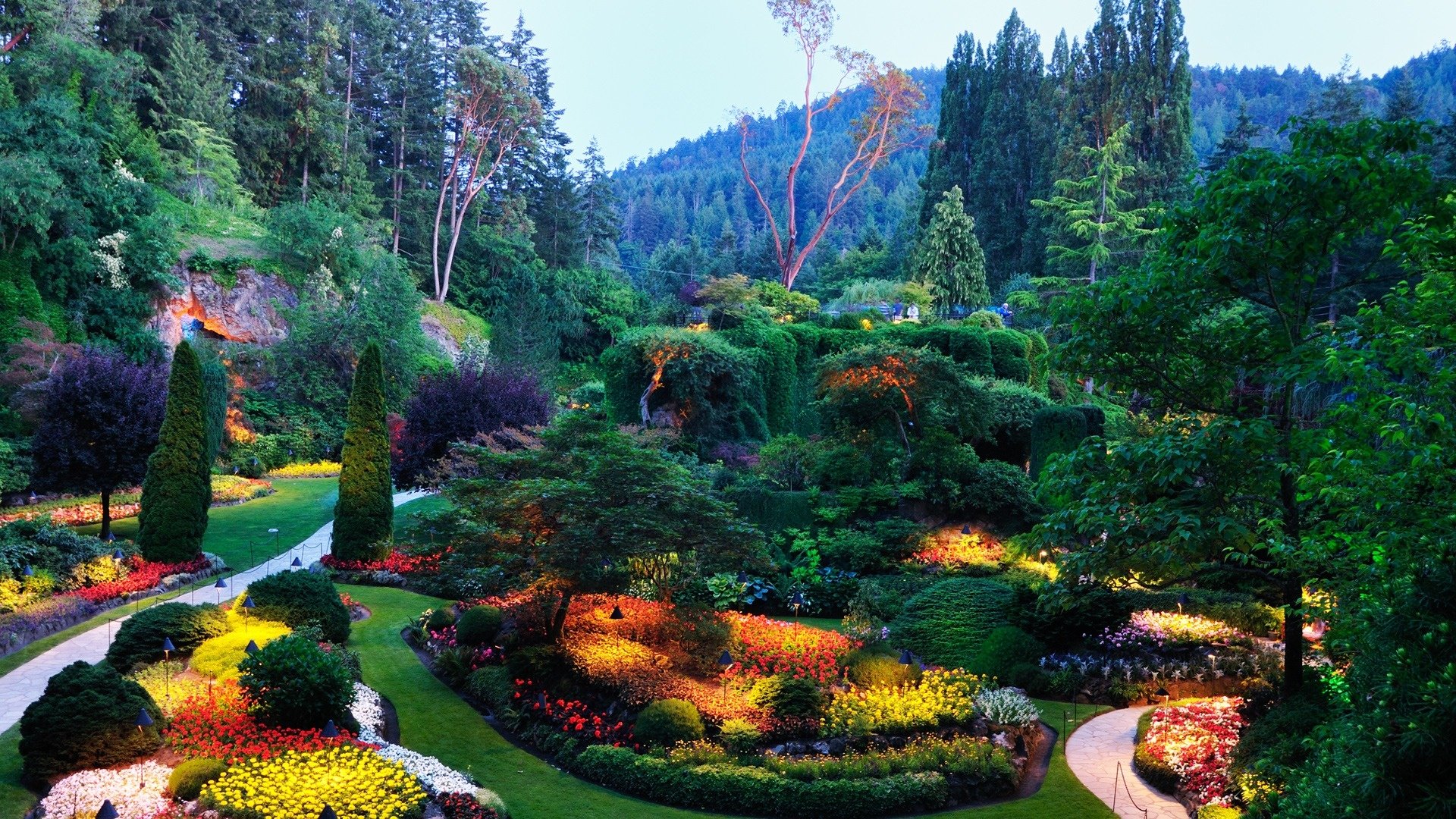 A lush, colorful garden with meticulously arranged flower beds and trees, set against a backdrop of rolling hills, creating a vibrant atmosphere for a HD desktop wallpaper.