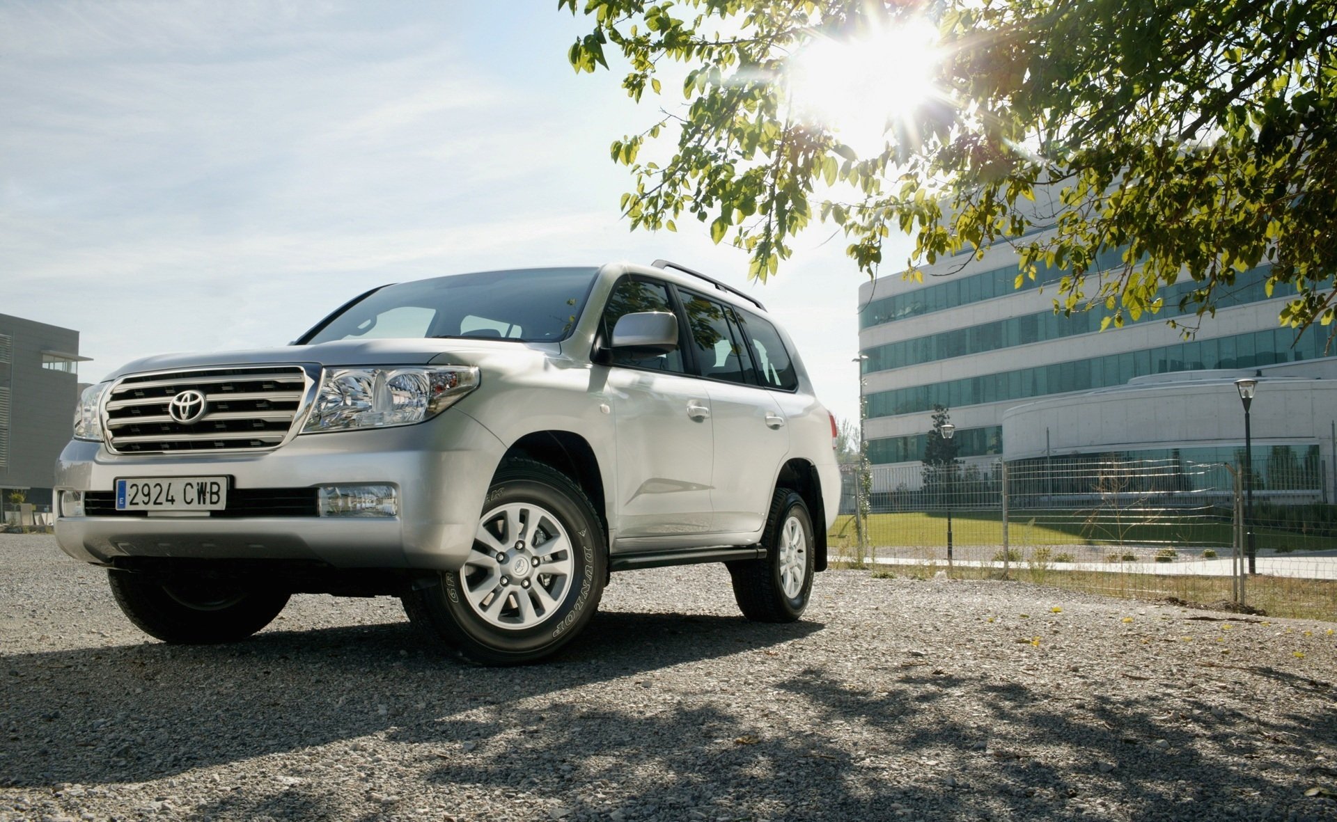 HD PC desktop wallpaper background featuring a silver Toyota SUV parked by a modern office building with sunlit trees overhead.