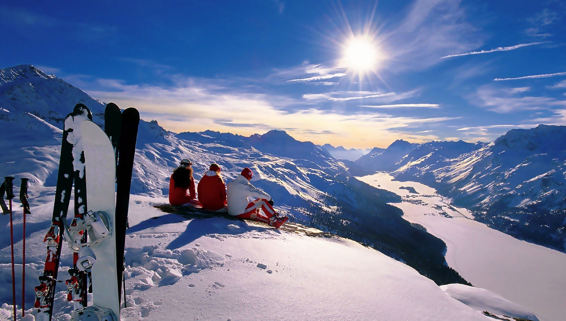 Photography HD PC desktop wallpaper: two people in red jackets sit on a snowy peak beside skis, gazing over a sunlit alpine valley and frozen lake under a bright blue sky.