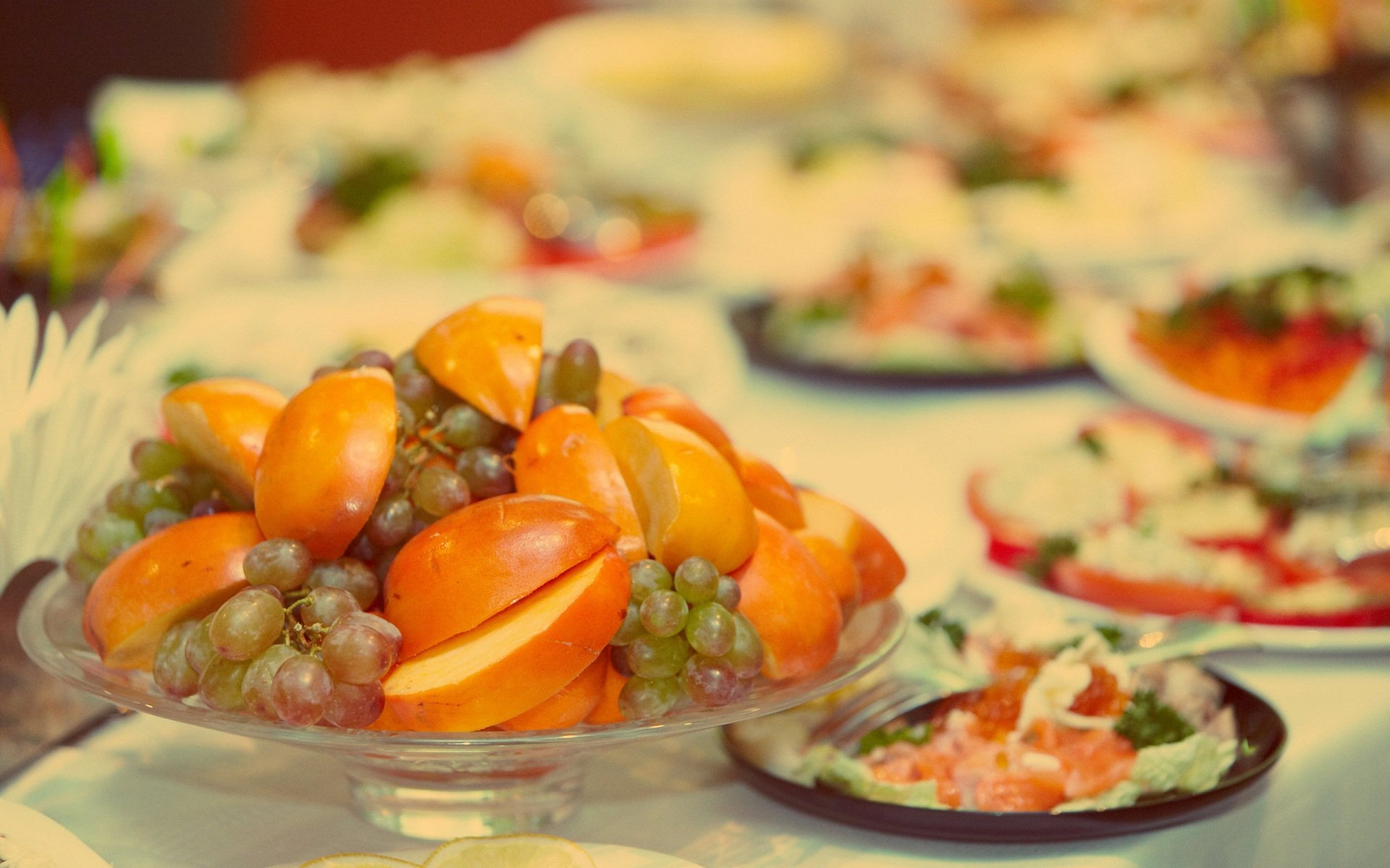 HD desktop wallpaper featuring fresh grapes and mango fruit artfully arranged on a table with plates of colorful food in the background.