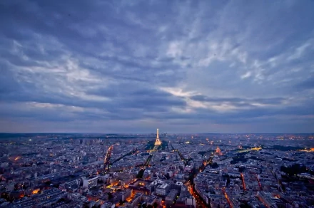 Aerial view of Paris at dusk with the illuminated Eiffel Tower under dramatic clouds, city lights stretching to the horizon — man-made urban panorama, 2K Quad HD PC desktop wallpaper/background.