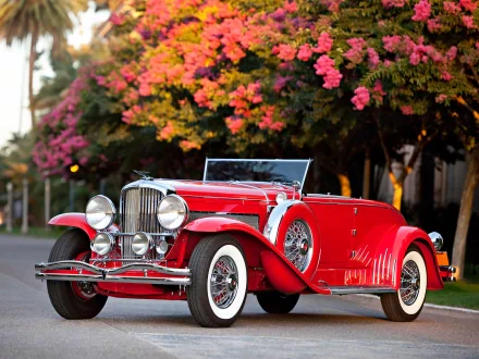 A striking red Duesenberg Model J parked on a scenic street, framed by vibrant pink flowers, showcasing its classic design and elegance in this HD desktop wallpaper.