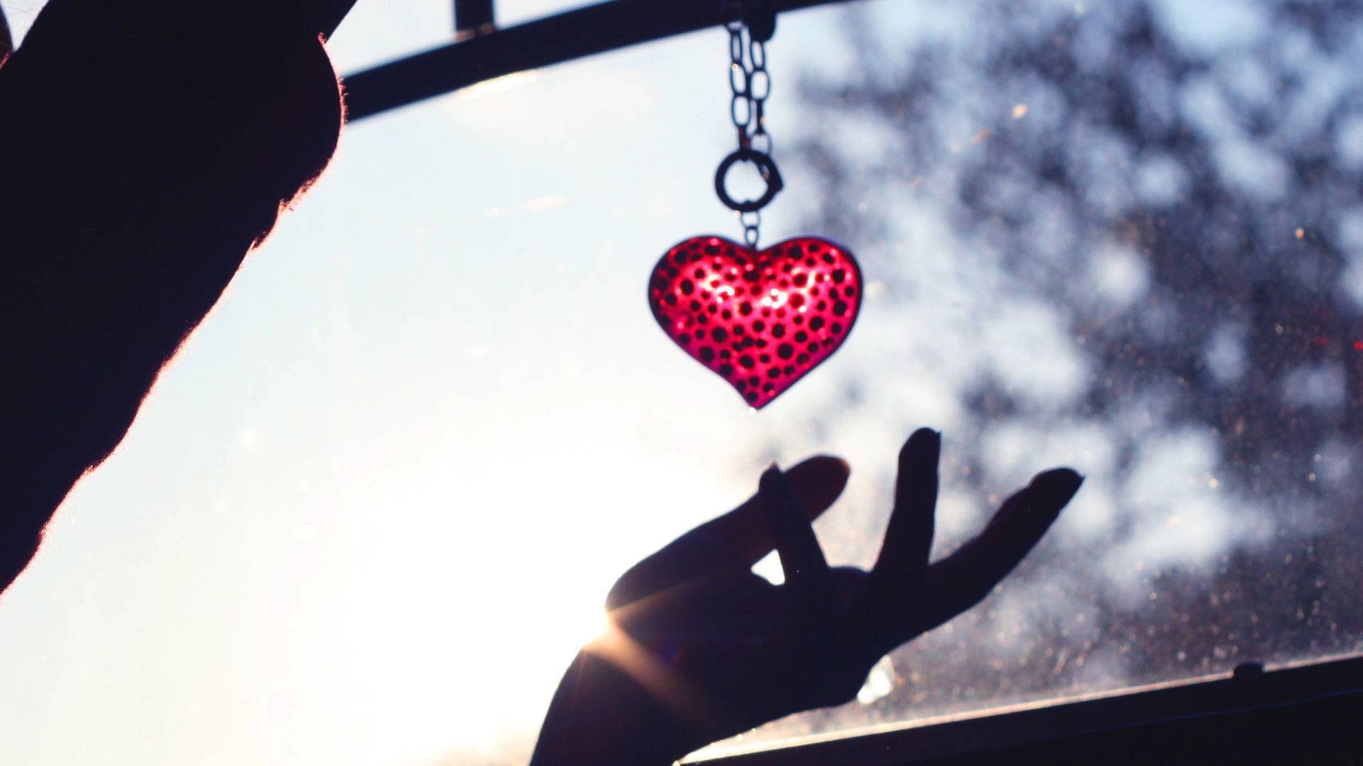 Artistic HD PC desktop wallpaper featuring a red heart pendant hanging above an outstretched hand, backlit by sunlight with a blurred natural background.