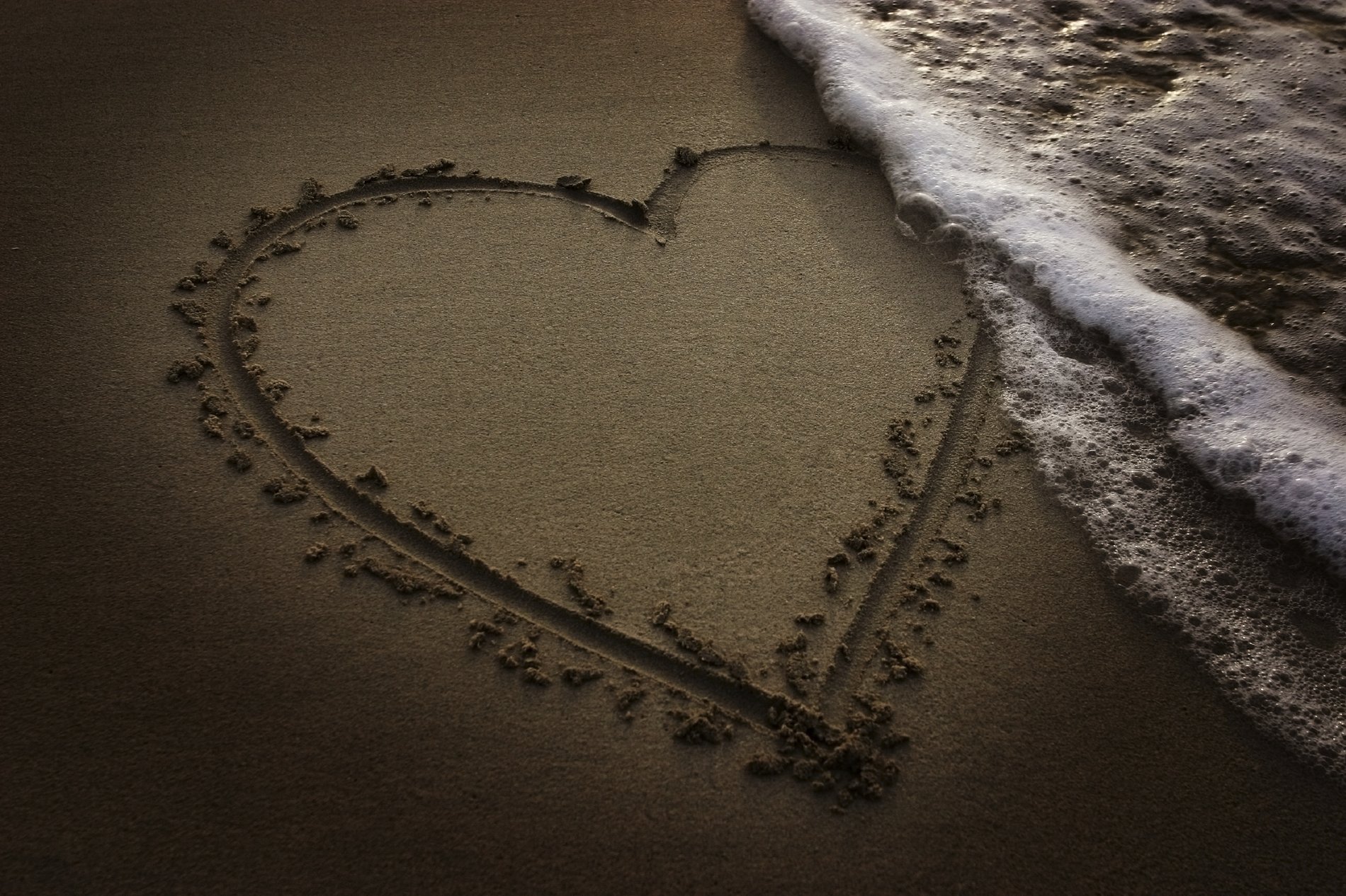 A heart drawn in sand with ocean waves gently washing over it. This HD desktop wallpaper and background conveys themes of love and artistic expression.