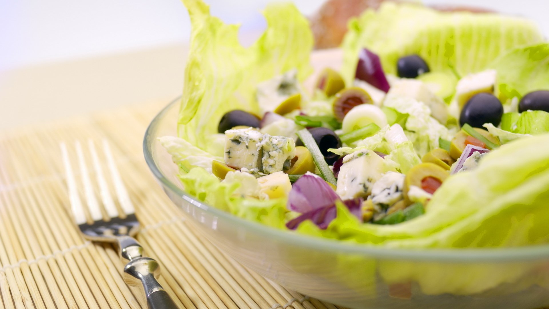 HD desktop wallpaper featuring a fresh salad with lettuce, olives, nuts, and herbs, presented in a clear glass bowl on a bamboo mat with a fork beside it.