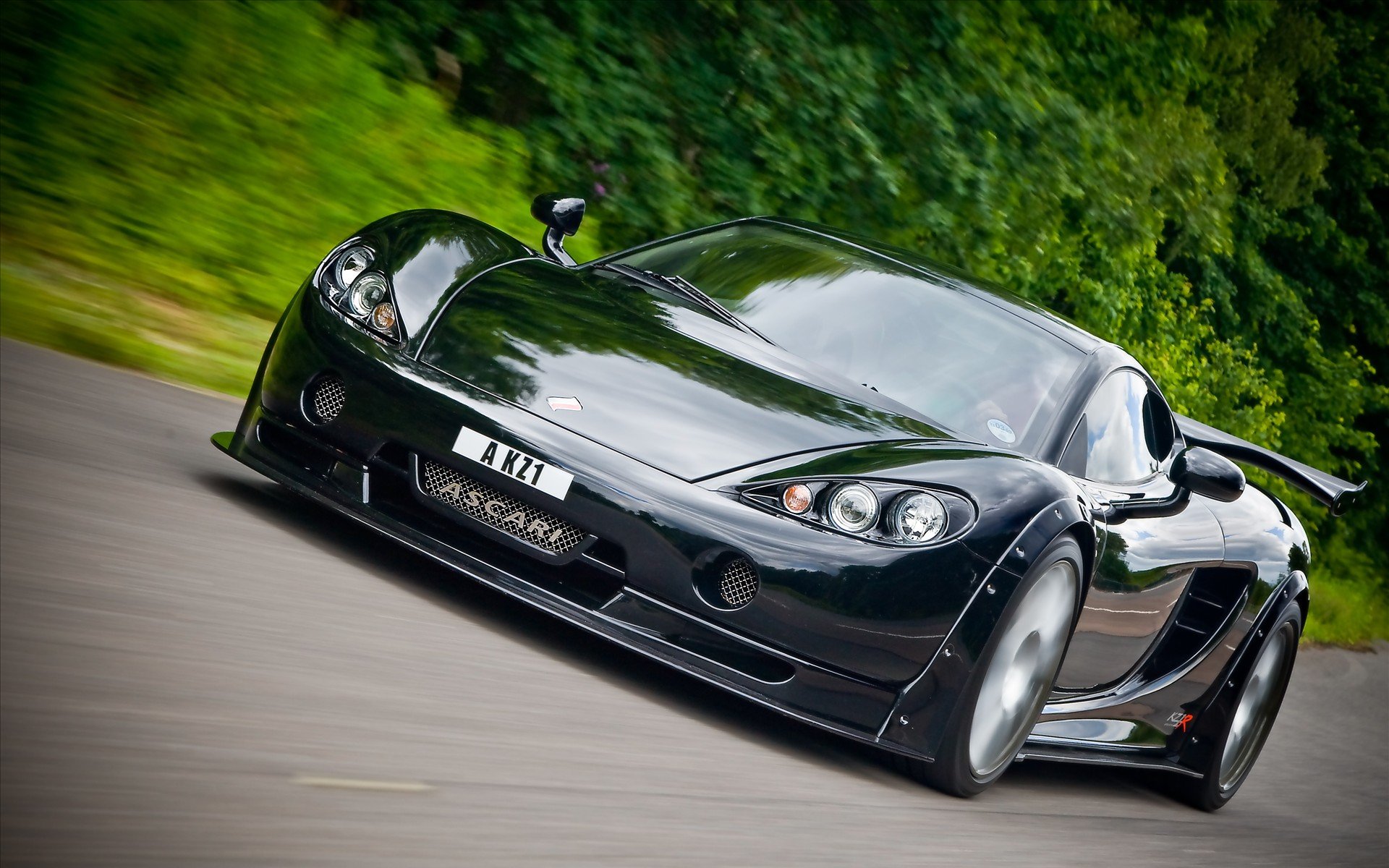 Sleek black Ascari KZ1 sports car racing on a tree-lined road, HD PC desktop wallpaper background showcasing the vehicle in motion.