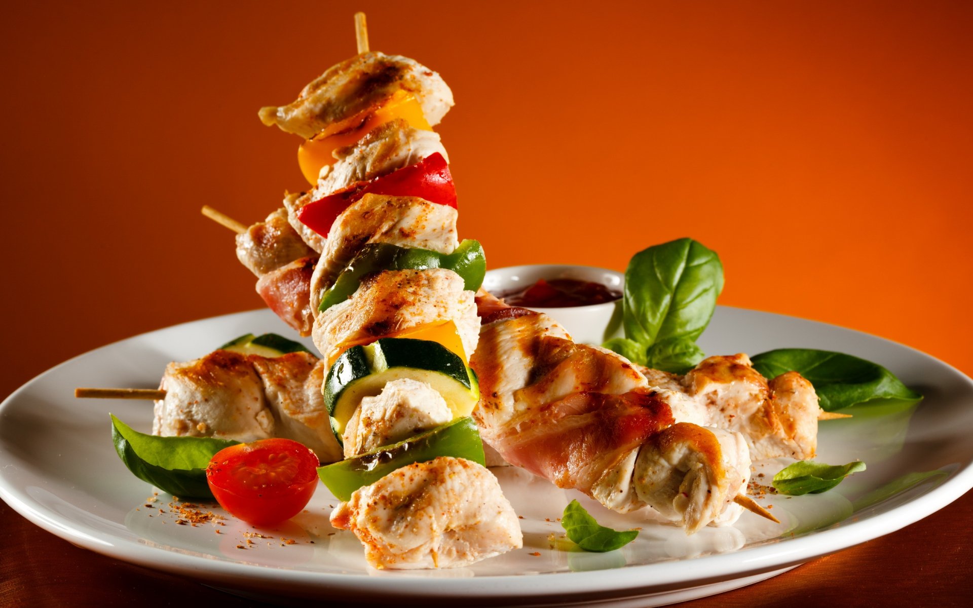 HD PC desktop wallpaper featuring colorful barbecue skewers with grilled chicken, vegetables, and fresh basil leaves on a white plate against a warm orange background.