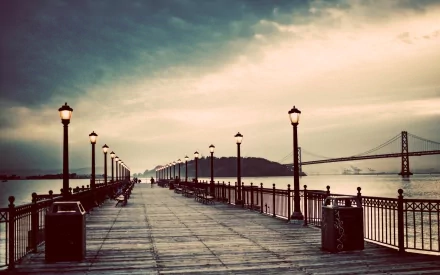 View of Pier 7 extending into the water with street lamps lining the wooden walkway and the Bay Bridge visible in the background under a cloudy sky, San Francisco.
