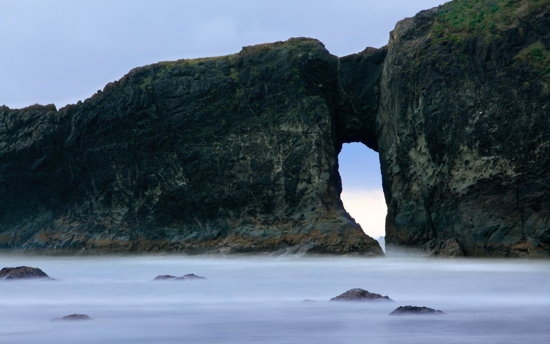 HD desktop wallpaper featuring a natural rock arch rising from misty waters against a soft blue sky, blending nature's rugged beauty with serene coastal elements.