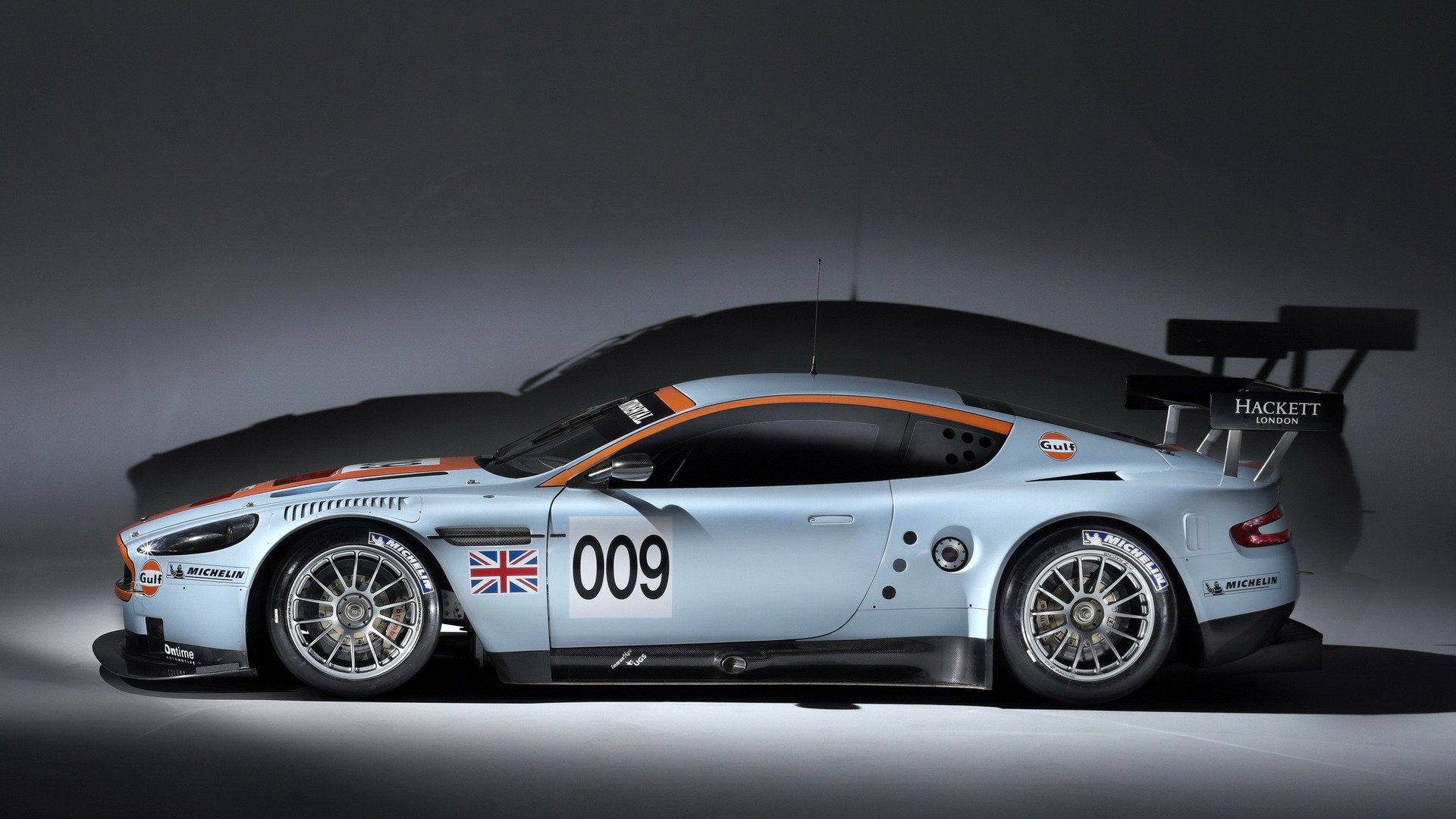 HD PC desktop wallpaper and background showing a silver racing sports car vehicle (side profile) marked 009 with British flag, aerodynamic body and rear wing on a neutral studio backdrop.