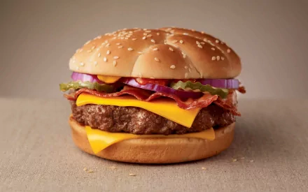 HD desktop wallpaper featuring a close-up of a juicy cheeseburger with bacon, lettuce, tomato, and onions on a sesame seed bun.
