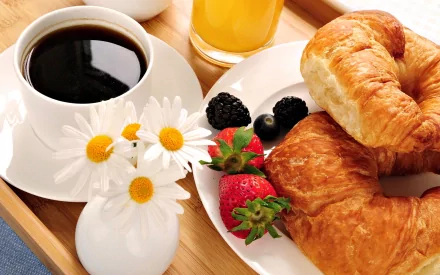 HD desktop wallpaper featuring a breakfast still life with coffee cup, orange juice, croissants, fresh fruit, and a small vase of daisies on a wooden tray.