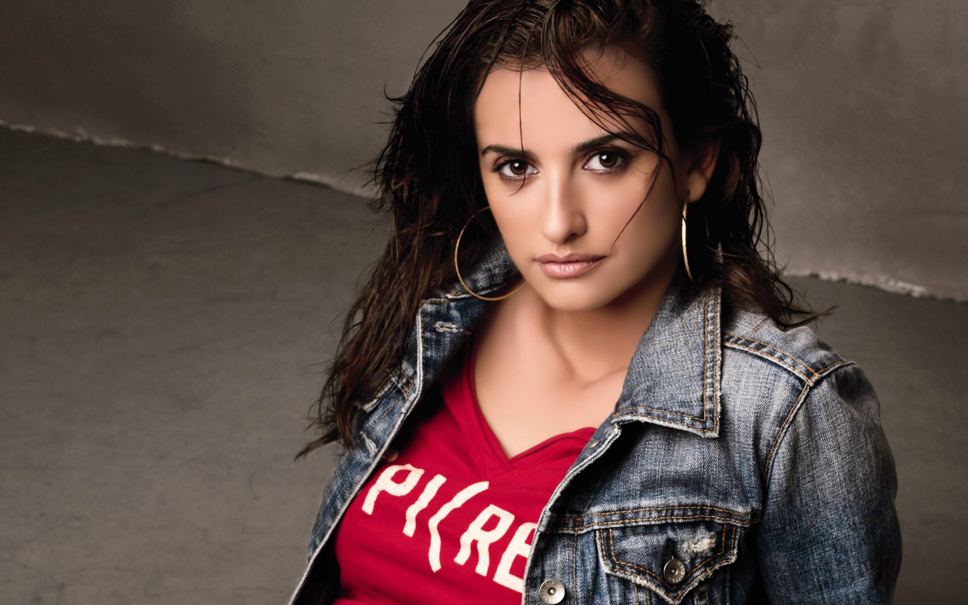 HD PC desktop wallpaper featuring celebrity Penelope Cruz in a casual denim jacket and red shirt, posing with a confident expression against a neutral background.