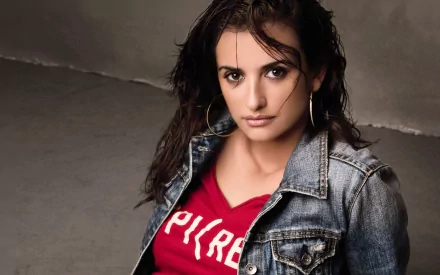 HD PC desktop wallpaper featuring celebrity Penelope Cruz in a casual denim jacket and red shirt, posing with a confident expression against a neutral background.