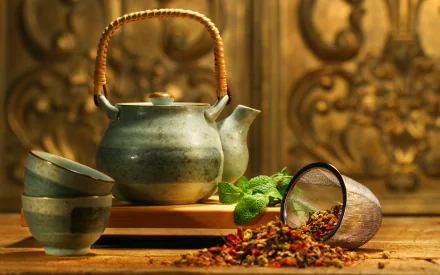 HD desktop wallpaper featuring a rustic teapot with stacked cups, spilled loose tea leaves, and fresh mint leaves on a wooden surface with an ornate background.