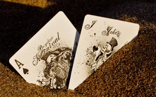 Close-up of two intricately designed playing cards, the Ace of Spades and a Joker, partially buried in sandy ground. This high-definition image serves as a striking desktop wallpaper.