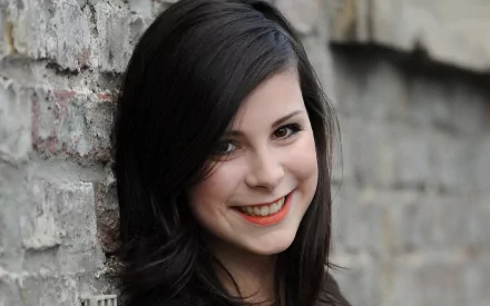 A close-up image of Lena Meyer-Landrut, showcasing her smiling against a textured brick wall, embodying the essence of music in this HD desktop wallpaper.