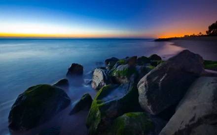 2K Quad HD PC desktop wallpaper and background: coastal nature scene with moss-covered rock formations in foreground, silky long-exposure water and a vivid sunset-to-blue gradient sky.
