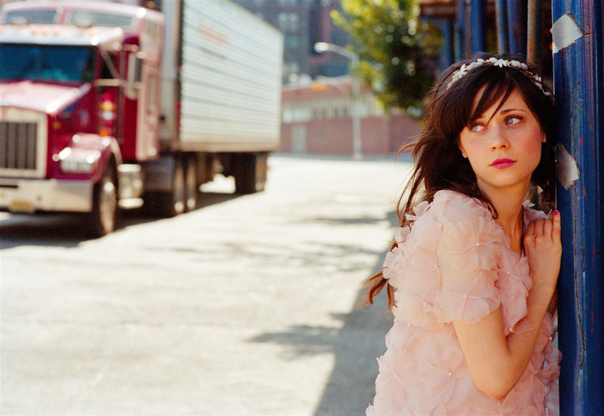 A captivating HD wallpaper featuring Zooey Deschanel, showcasing her in a soft, pink outfit against an urban backdrop with a large truck in the background.