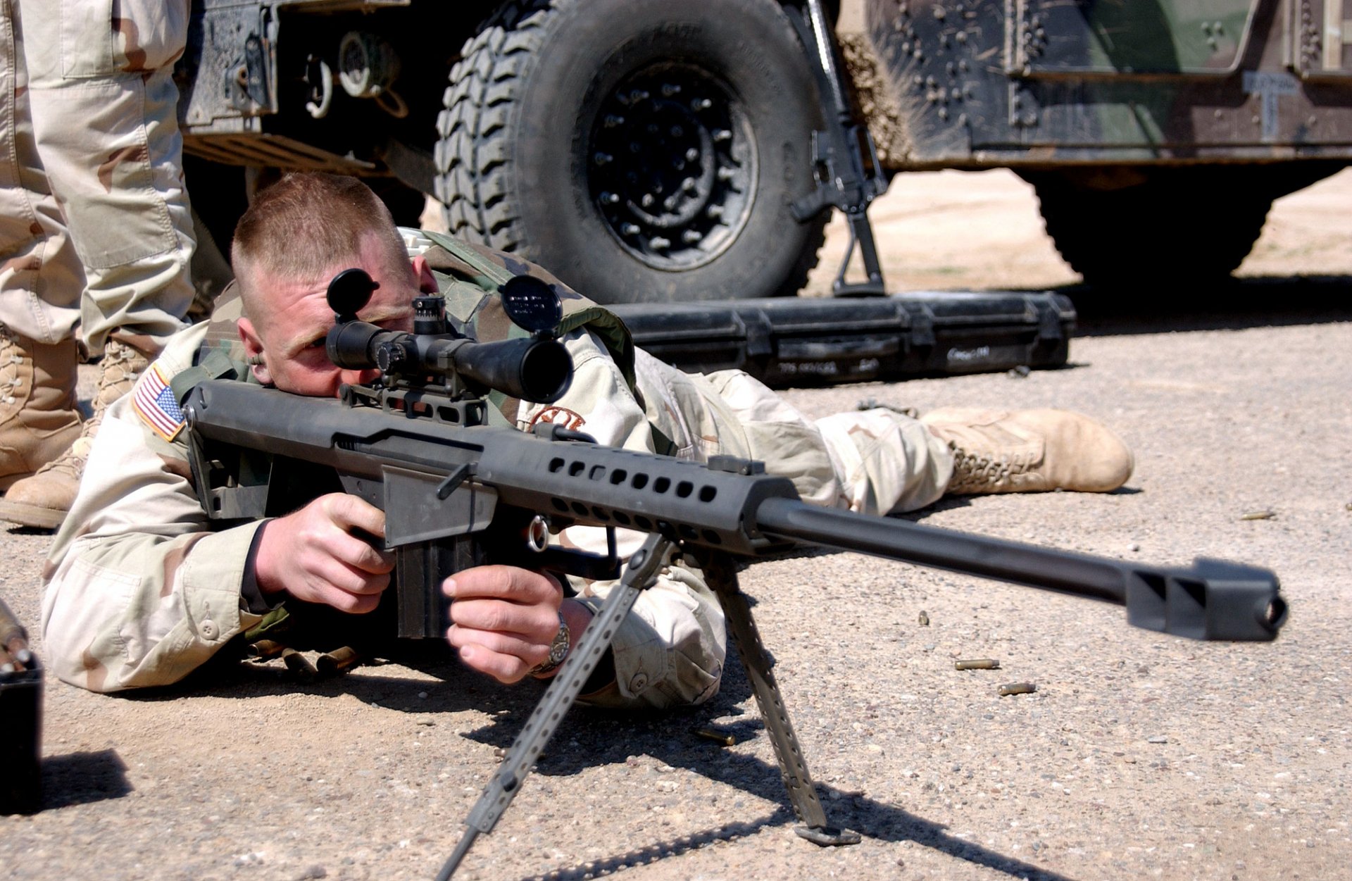 A military sniper aims a .50 cal sniper rifle while lying on the ground, with military vehicles in the background. HD desktop wallpaper and background.