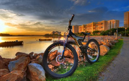 A stunning HD wallpaper featuring a bicycle resting near a serene waterfront at sunset, surrounded by rocks and apartment buildings, showcasing a perfect blend of nature and urban life.