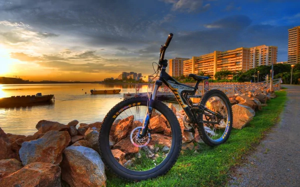 A stunning HD wallpaper featuring a bicycle resting near a serene waterfront at sunset, surrounded by rocks and apartment buildings, showcasing a perfect blend of nature and urban life.