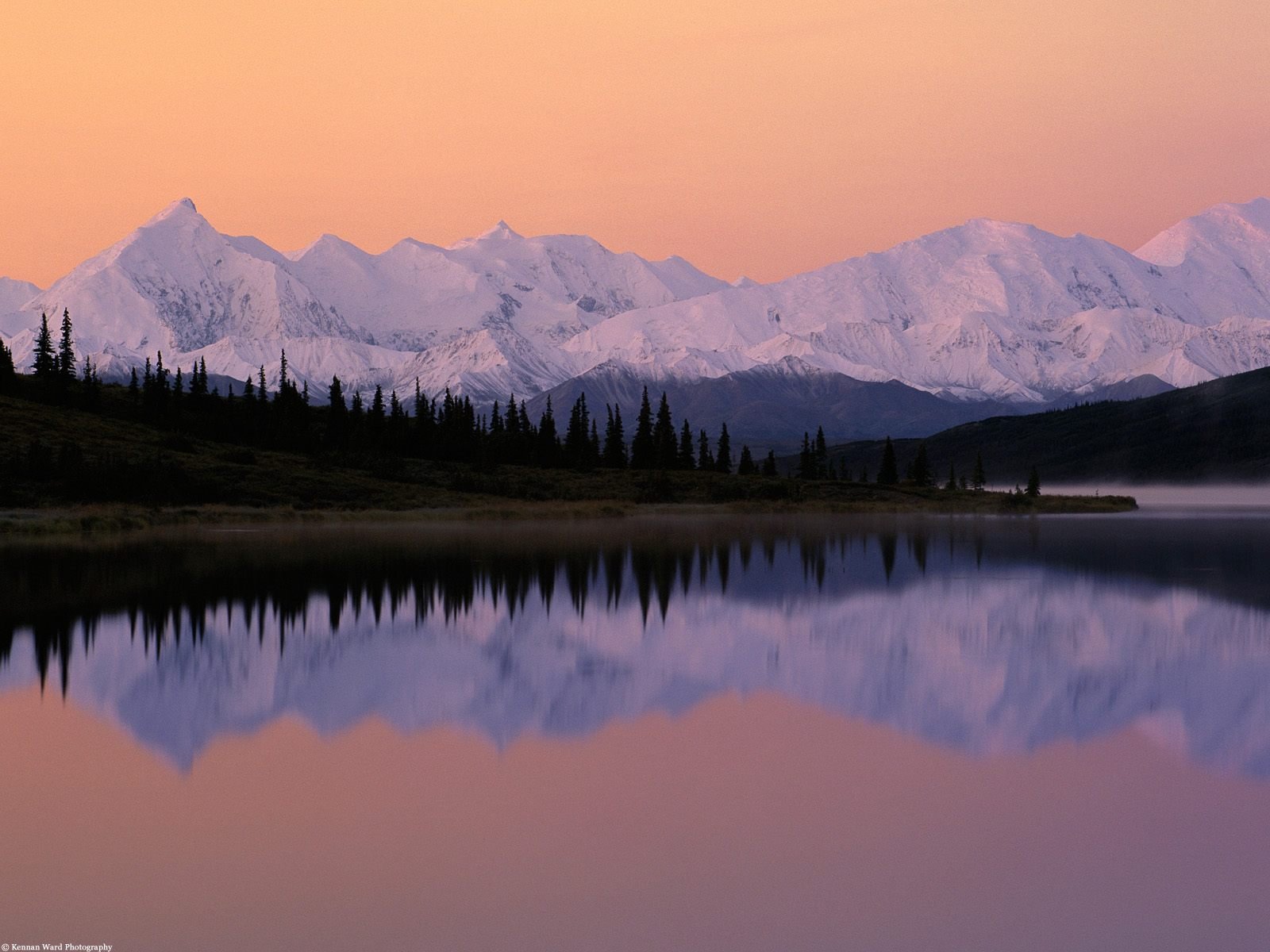 Snow-capped mountains rise majestically above a tranquil lake, their reflection shimmering in the water under a soft, colorful sky. A breathtaking nature scene for any HD desktop background.