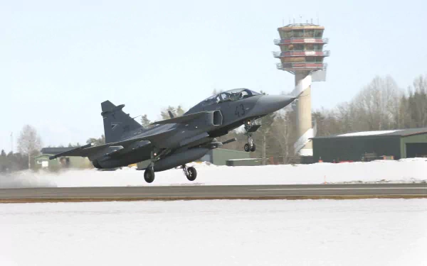 A Saab JAS 39 Gripen military jet takes off against a snowy backdrop, with an air traffic control tower visible in the background, creating a striking HD desktop wallpaper.