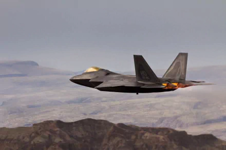 HD desktop wallpaper of a Lockheed Martin F-22 Raptor military jet flying over rugged terrain under a cloudy sky.