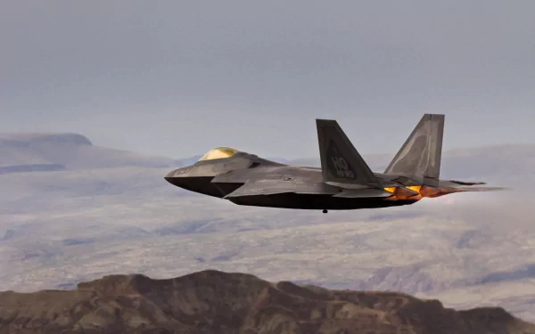 HD desktop wallpaper of a Lockheed Martin F-22 Raptor military jet flying over rugged terrain under a cloudy sky.