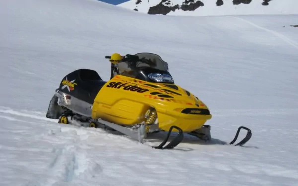 Yellow snowmobile vehicle on a snowy mountain slope beneath rocky peaks — HD PC desktop wallpaper background.