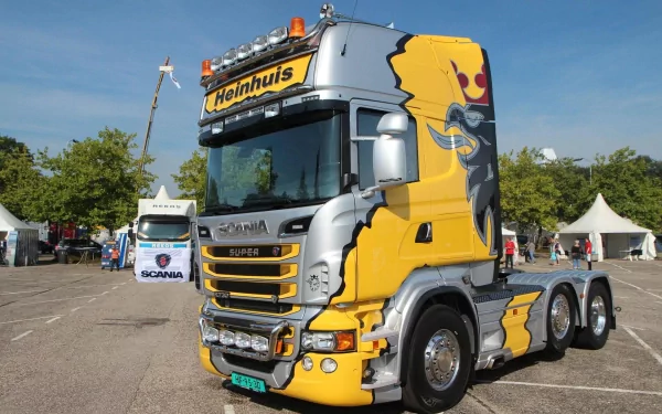 A vibrant Scania truck with bold graphics, prominently displaying the Heinhuis logo, parked at an event with tents and trees in the background, showcased as HD desktop wallpaper.