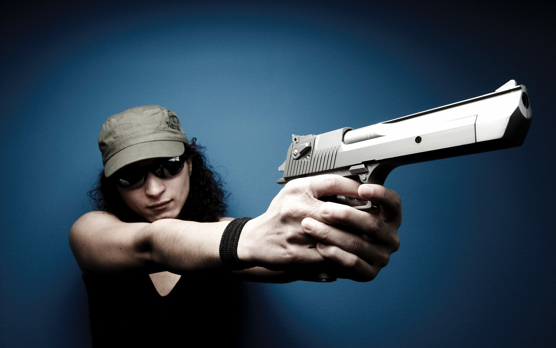 HD PC desktop wallpaper of a woman wearing sunglasses and a cap, aiming a silver handgun against a dark blue background, tagged Girls & Guns.