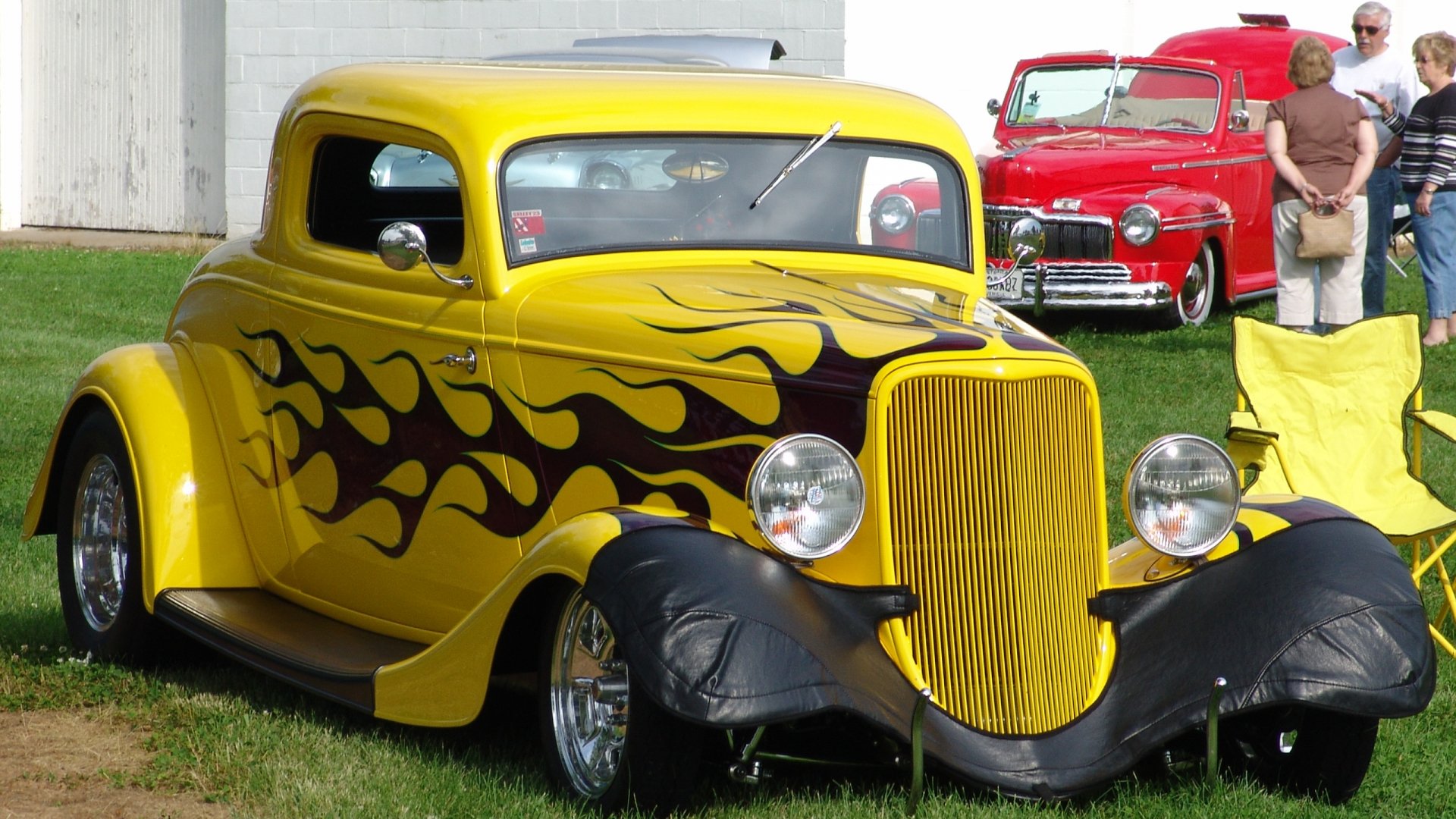 HD PC desktop wallpaper featuring a vintage yellow car with black flames and a classic red truck in the background at an outdoor event.