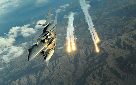 A McDonnell Douglas F-15E Strike Eagle maneuvers through the sky, releasing flares over rugged terrain, showcasing its military capability as a powerful jet fighter.