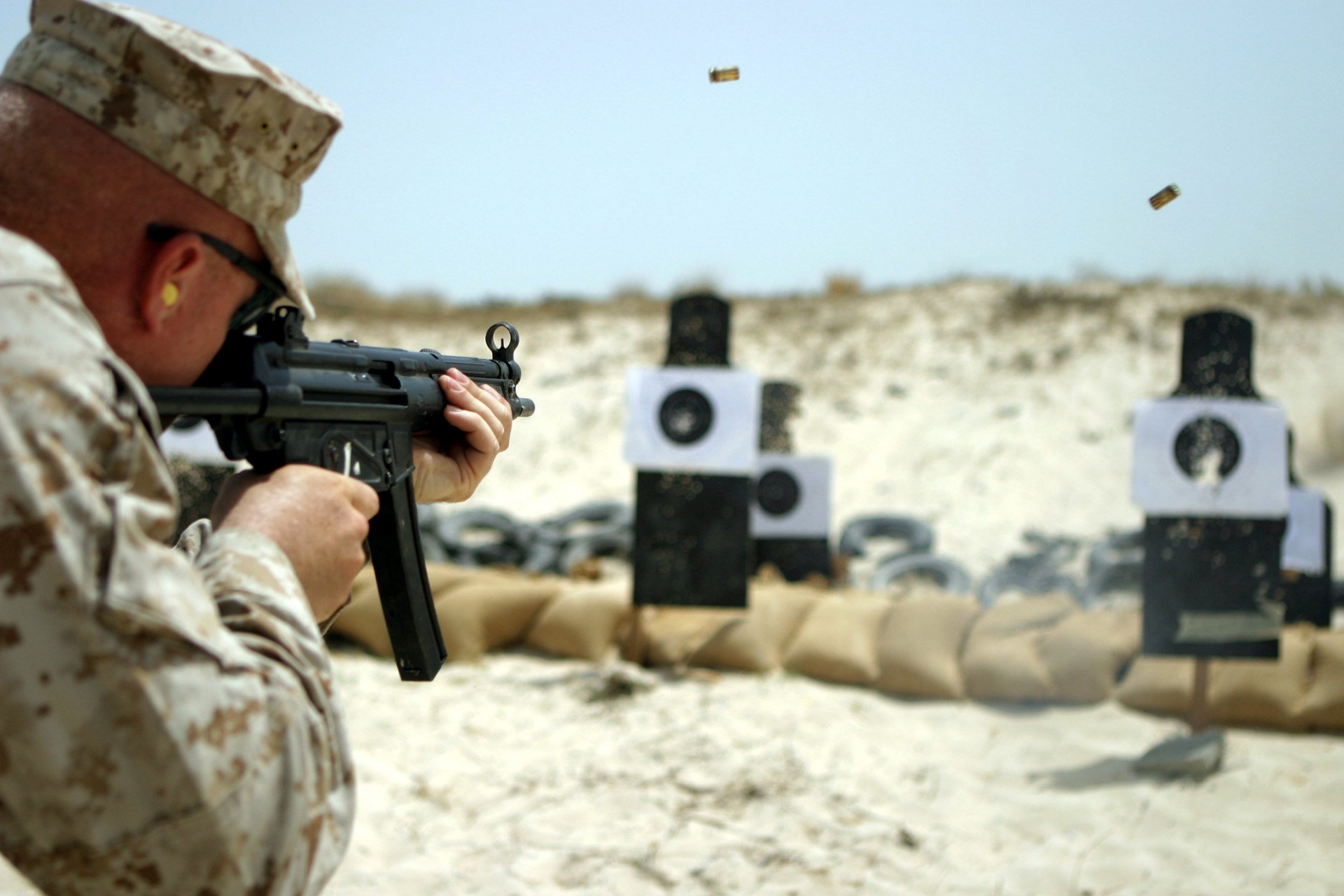 HD desktop wallpaper showing a soldier in military gear aiming a gun at paper targets on a sandy outdoor firing range.
