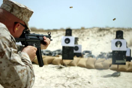 HD desktop wallpaper showing a soldier in military gear aiming a gun at paper targets on a sandy outdoor firing range.